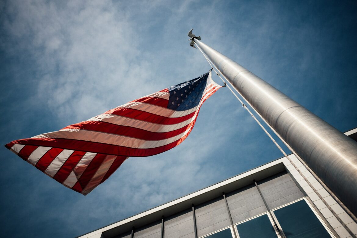 a bottom up view of an american flag attached to a tall silver pole flying in the.wind in front of a grey office building with windows