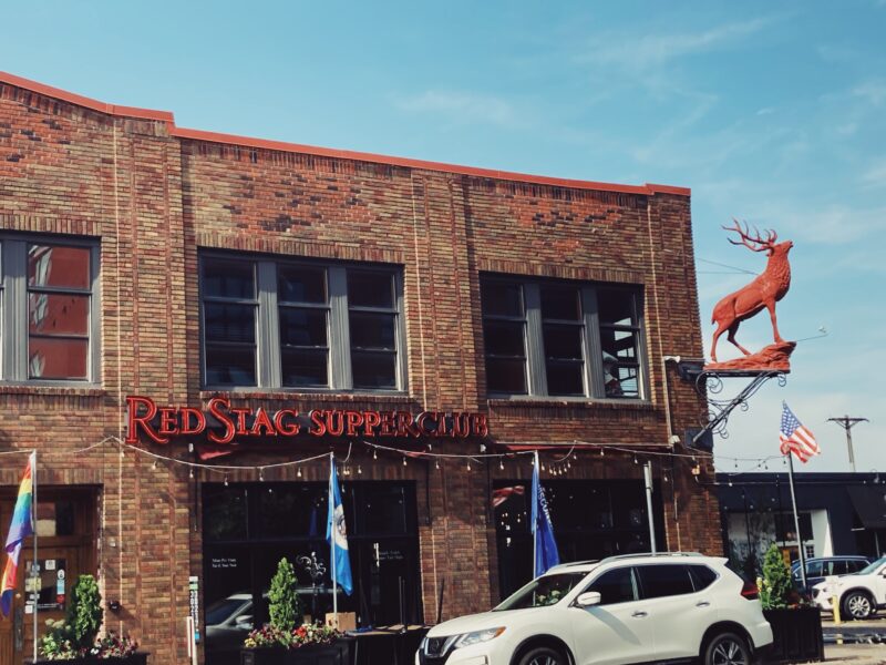 the storefront of the Red Stag Supperclub during the daytime. which has its name in big red letters and a big red stag mannequin hanging off the corner of the brick building