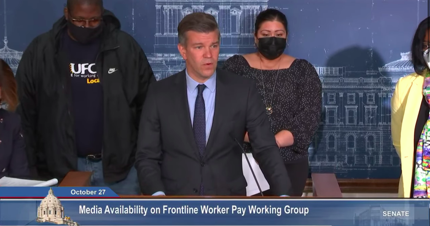 screenshot of press conference man in dark suit and blue shirt standing behind a podium in front of two people one wearing a black sweatshirt and other wearing a black dress and black face mask