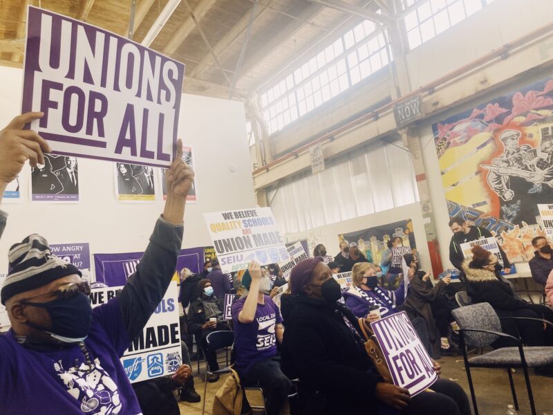 a group of workers wearing purple tee shirts hold up signs, one worker toward the front left holds up a sign saying "unions for all"