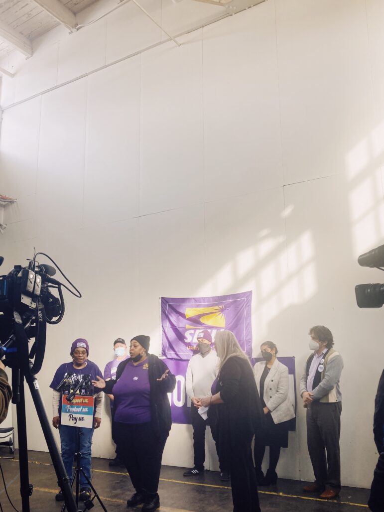 video cameras frame a group of people standing in front of a purple SEIU poster in front of a microphone