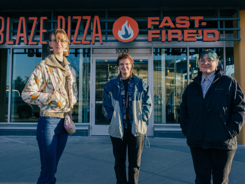 three people stand in front of the blaze pizza storefront, one wears a khaki jacket, one a denim jacket, and one a black jacket with blue hair
