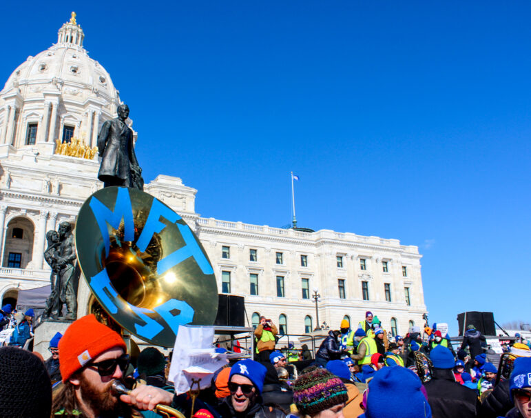 a tuba player holding a tuba that has blue painted letters of MFT ESP plays in front of a backdrop of the state capitol building underneath a clear blue sky