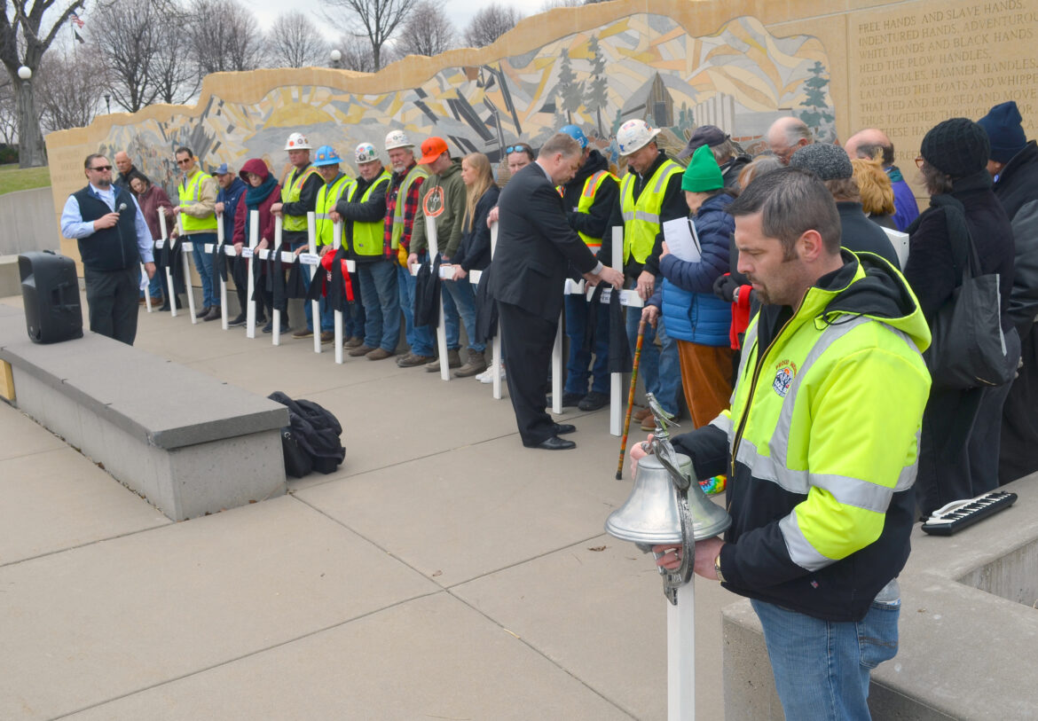 a man in a neon yellow construction jacket holds a silver bell, while people, some wearing neon yellow vests and white hard hats, line up in front of the worker's memorial with 13 white crosses