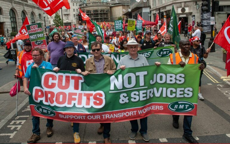 the front of a crowd of protestors marching hold a green and red banner reading "Cut Profits not jobs & services" and "support our transport workers"