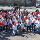 A group of workers and union members wearing white shirts and red hats stand in a large crowd with their fists raised
