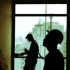 Two silhouettes of men standing in front of a window wearing hard hats and handling construction gear