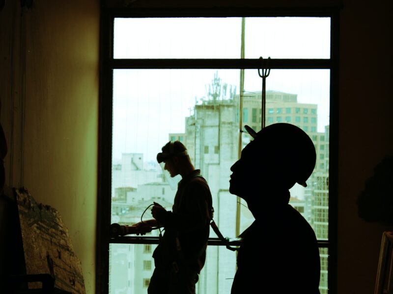 Two silhouettes of men standing in front of a window wearing hard hats and handling construction gear