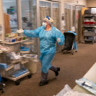 a healthcare worker wearing a blue PPE suit and white mask, gloves, and face shield rushes down a hospital hallway reaching out a hand to a hand sanitizer. the background is slightly blurred to convey movement