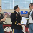 a man in a decorated military outfit with a beret stands between a woman with a US flag in her hand and a man with a blazer on, holding a white paper plaque
