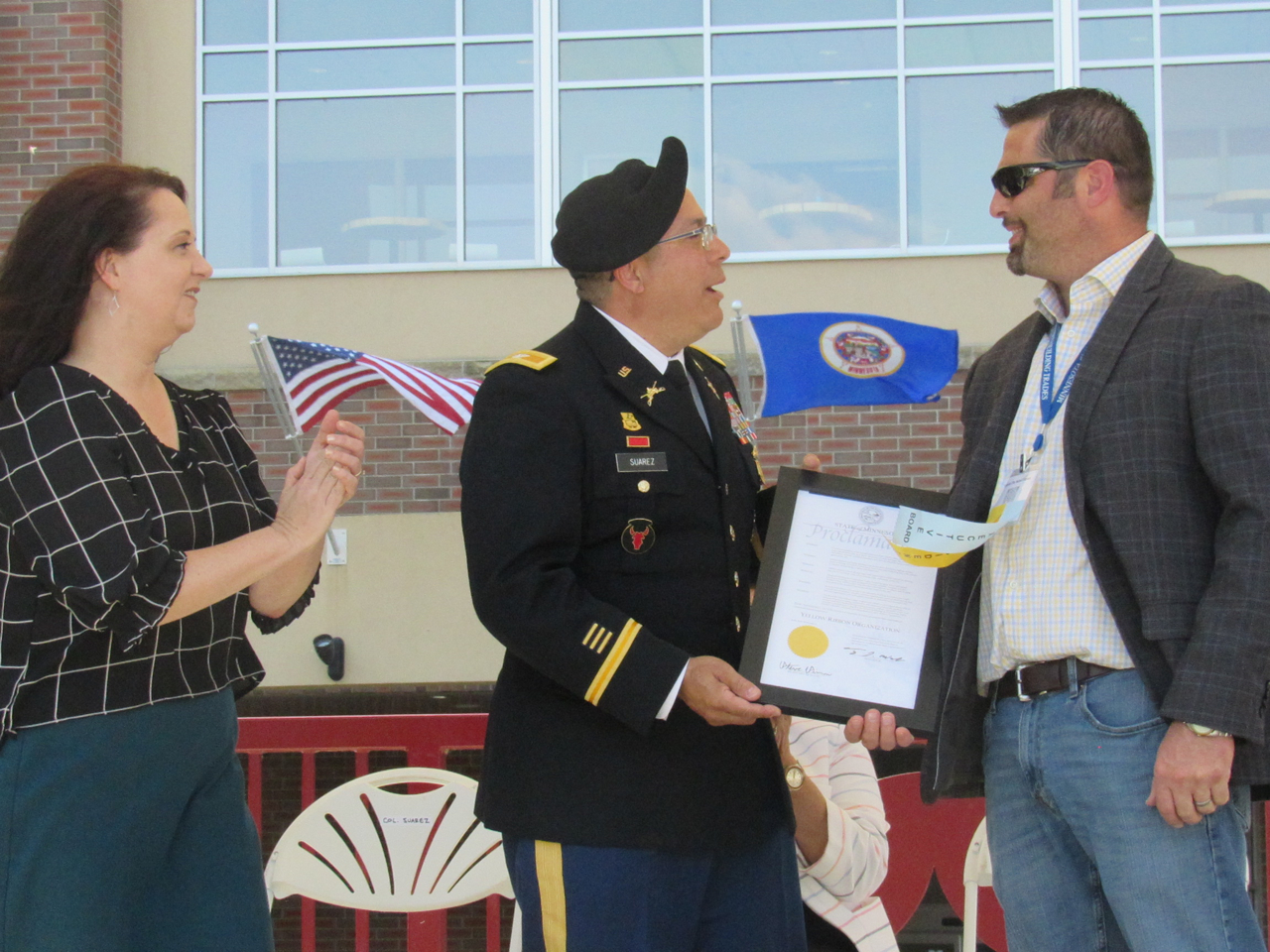 a man in a decorated military outfit with a beret stands between a woman with a US flag in her hand and a man with a blazer on, holding a white paper plaque