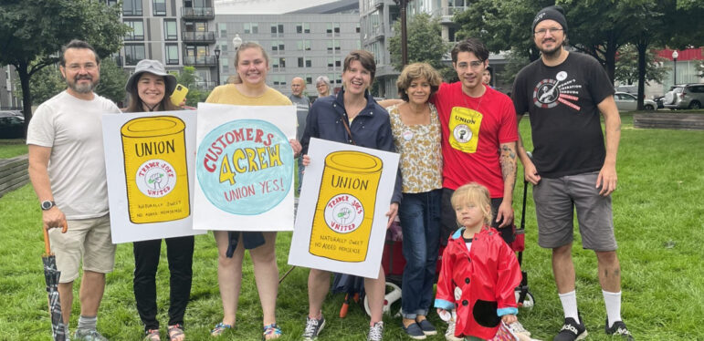 7 adults and one child stand in a row smiling holding blue, yellow, and white signs depicting a trader joe's soup can that says "union"