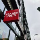 looking up at a red shop sign that reads "trader joe's" in white, a lampost and building are in the background