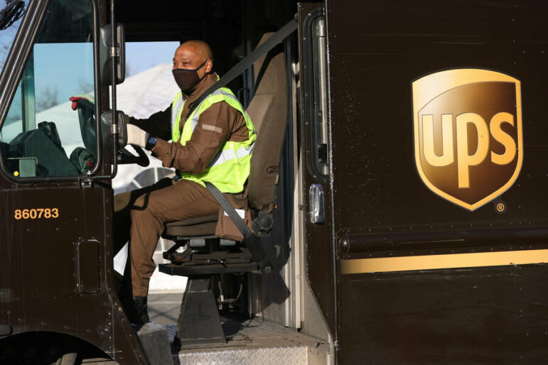A Black man wearing a black mask and a neon yellow vest sits in the driver's seat of a brown UPS truck with his hands on the wheel. The brown and yellow UPS logo is on the right side of the image of the truck