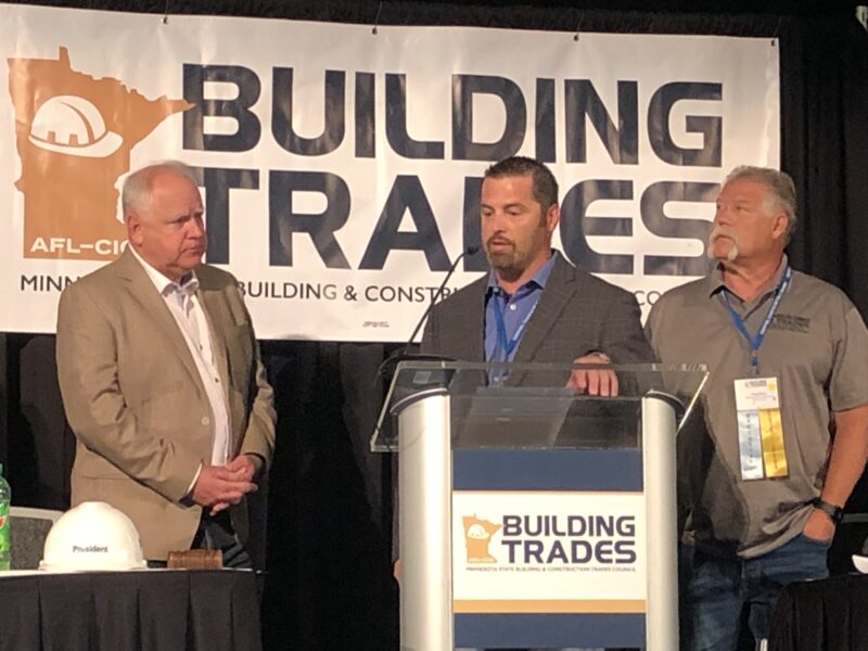 A man with a grey blazer stands at a podium between a man with a grey shirt and a man with a tan blazer in front of a sign that reads "BUILDING TRADES" and a logo of the state of Minnesota with a white hard hat
